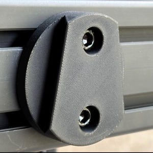 May include: Close-up of a dark gray, textured plastic component with two recessed screw holes. The component is attached to a series of parallel, gray metal bars. The design is functional and industrial.