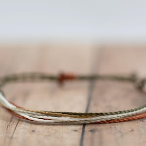 May include: A braided bracelet featuring strands of white, tan, olive green, and rust-colored threads. The bracelet is knotted at the ends and rests on a light brown wooden surface. The bracelet is a simple, minimalist design.
