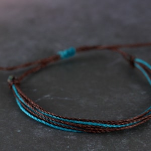 May include: A brown and teal braided bracelet with a sliding knot closure.