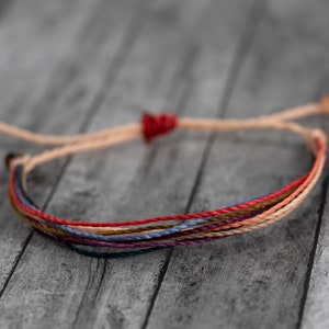 May include: A multi-colored friendship bracelet with red, orange, yellow, green, blue, and purple threads. The bracelet is tied with a knot at the end.