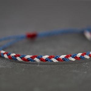 May include: A braided friendship bracelet with red, white, and blue threads. The bracelet is tied with a slip knot.