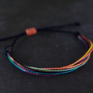 May include: A multi-colored friendship bracelet with a dark blue cord and a knot at the end. The bracelet is made of several strands of colorful thread, including pink, orange, yellow, green, and blue.