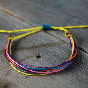 May include: A multi-colored friendship bracelet with a yellow cord and a turquoise bead. The bracelet is made of several strands of different colored cords, including pink, orange, red, purple, blue, and gray.
