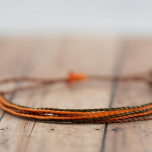 May include: A handmade bracelet featuring three strands of cord in shades of brown, orange, and olive green. The bracelet is knotted at the ends and has an adjustable closure. The bracelet is displayed on a wooden surface.