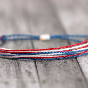 May include: A red, white, and blue friendship bracelet made of woven string. The bracelet is tied with a knot and has a small white bead.