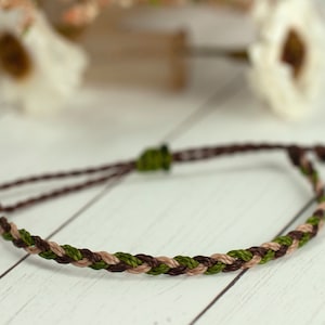 May include: Braided bracelet featuring brown, green, and tan threads. The bracelet has an adjustable closure with brown cords and a small green accent. The bracelet is displayed on a white wooden surface.