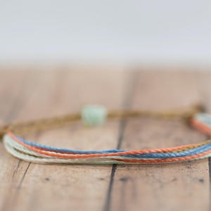 May include: A multi-colored friendship bracelet with a light green bead. The bracelet is made of woven string in shades of blue, peach, and white.