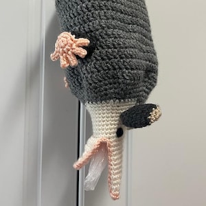 Puede incluir: Una bolsa hecha a mano con forma de zarigüeya de ganchillo, principalmente gris con una cara crema y detalles en rosa. La bolsa tiene un ojo negro y una pequeña bolsa de plástico que sale de la boca. La zarigüeya tiene pequeñas manos rosas de ganchillo.