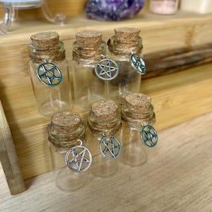 May include: Six small, clear glass bottles with cork stoppers and silver pentacle charms. The bottles are arranged on a wooden shelf. The bottles are empty, ready to be filled, suggesting a theme of witchcraft or spiritual practices.