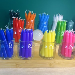May include: A selection of brightly coloured spell candles displayed in glass jars. The candles are in shades of blue, red, orange, green, and pink. The jars are arranged on a wooden tray, against a green backdrop.