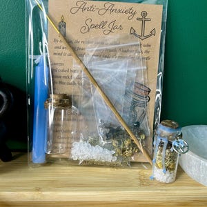 May include: An "Anti-Anxiety Spell Jar" kit, packaged in a clear plastic bag. The kit includes a blue candle, a small glass bottle with a cork, an incense stick, and a card with text and an anchor illustration. The jar contains crystals and herbs.