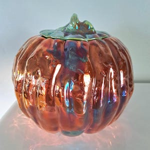 May include: A ceramic pumpkin-shaped jar with a lid. The jar has a glossy, iridescent finish in shades of orange, red, and blue. The lid and stem are green. The pumpkin has a ribbed texture.