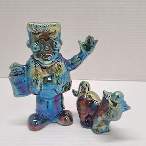 May include: A pair of iridescent blue and purple glass figurines featuring a standing character with a flat-top hairstyle holding a bucket labeled Trick or Treat and a smaller four-legged animal figure wearing a hat, both with a reflective finish.