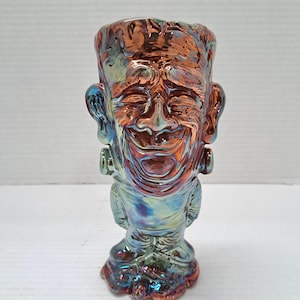 May include: A ceramic tiki mug shaped like a laughing Frankenstein monster with an iridescent, metallic copper and blue finish. The mug features a detailed facial expression, prominent bolts on the neck, and a textured surface throughout the design.