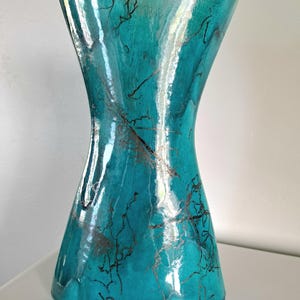 May include: A turquoise hourglass-shaped vase with a flared top and base. The glass vase has a glossy finish and is decorated with silver and black abstract lines. The vase is displayed on a white surface.
