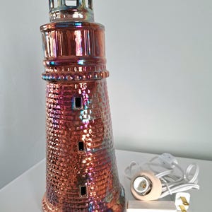 May include: A ceramic lighthouse lamp with a textured brick pattern and iridescent finish. The lamp has a copper and purple color scheme, with a small light fixture and cord. The lamp is approximately 12 inches tall.