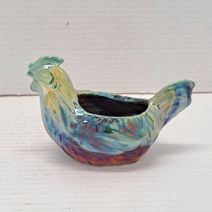 May include: A ceramic hen-shaped planter with a vibrant, iridescent glaze. The planter features a detailed head and tail, with a wide opening for planting. The colors include shades of blue, green, yellow, and red, creating a unique decorative piece.