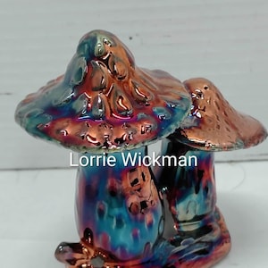 May include: A ceramic sculpture featuring two mushrooms with a metallic, iridescent glaze in shades of blue, purple, and copper. The text Lorrie Wickman is printed across the center of the mushrooms. The base is a textured, copper-colored surface.