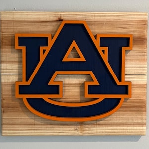 May include: A wooden sign with the letters "AU" in a navy blue color with an orange outline. The letters are raised and have a 3D effect.