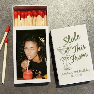 May include: A white matchbox with a photo of a woman winking and drinking from a straw. The matchbox cover reads "Stole This From" with a martini glass graphic and the text "Amelia's 21st Birthday 11.17.2025". Two matches are next to the box.
