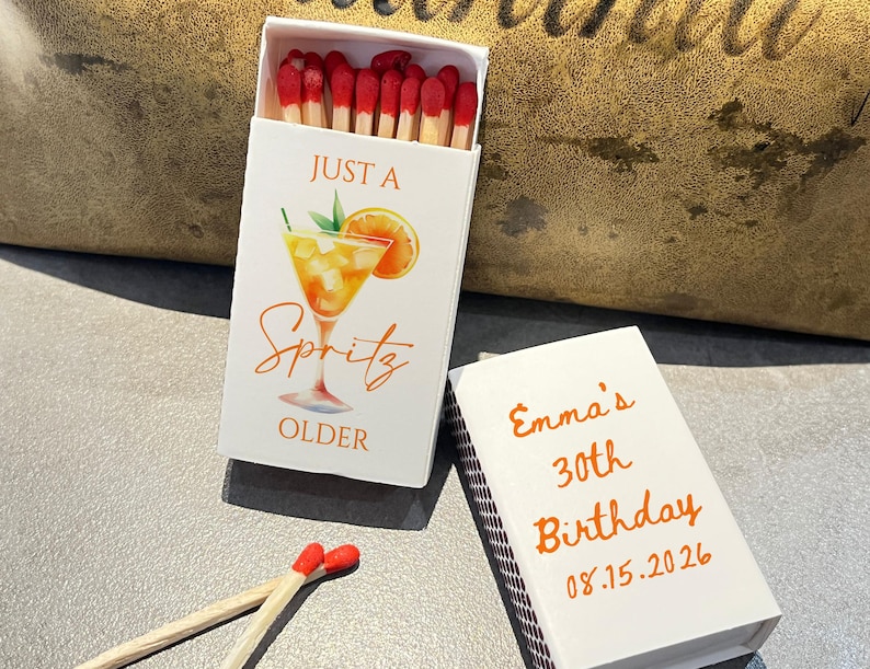 May include: White matchbox with orange text and a cocktail illustration that reads "Just a Spritz OLDER". Another matchbox reads "Emma's 30th Birthday 08.15.2026". Matches with red tips are visible.