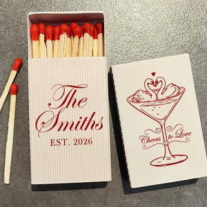 May include: A rectangular matchbox with a white and light gray pinstripe pattern, featuring the red text The Smiths EST. 2026. Beside it, a white matchbox cover displays a red illustration of two swans in a martini glass with the text Cheers to Love.