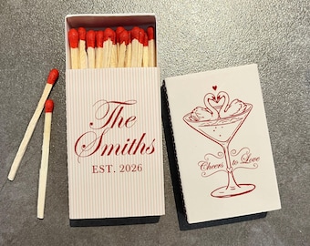 May include: A rectangular matchbox with a white and light gray pinstripe pattern, featuring the red text The Smiths EST. 2026. Beside it, a white matchbox cover displays a red illustration of two swans in a martini glass with the text Cheers to Love.