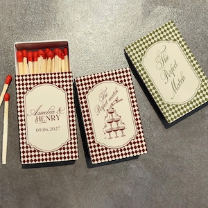 May include: Three rectangular matchboxes with a checkered pattern. One open box shows red-tipped matches. The boxes feature white labels with maroon text reading Amelia & Henry 09.06.2027, The Perfect Match with a champagne tower, and The Perfect Match.