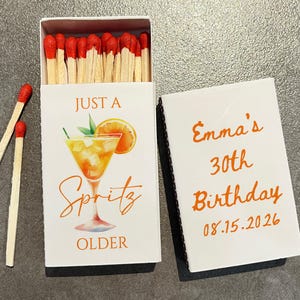 May include: A white matchbox with the text "JUST A Spritz OLDER" and a cocktail illustration. The open box reveals red-tipped matches. The adjacent matchbox cover reads "Emma's 30th Birthday 08.15.2026" in orange.