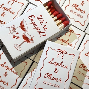 May include: White matchboxes with red text and illustrations. The text reads "Sophia & Oliver 02.15.2026" and "We're Engaged!". The match heads are red.