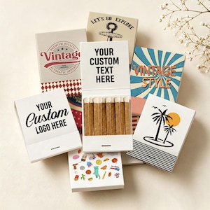 May include: A collection of rectangular matchboxes with various designs, including one open box revealing wooden matches with white tips. Text on the boxes includes "LET'S GO EXPLORE," "VINTAGE," "VINTAGE STYLE," "YOUR CUSTOM TEXT HERE," and "YOUR CUSTOM LOGO HERE."