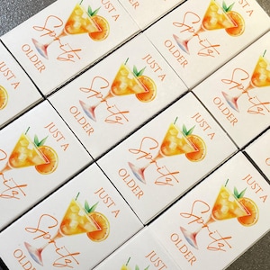 May include: White boxes with a watercolor illustration of a spritz cocktail, an orange slice, and text that reads "Just a Spritz Older". The boxes are arranged in a grid pattern.