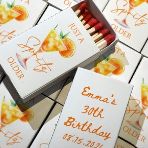 May include: White matchboxes with orange text and a watercolor illustration of a spritz cocktail. One box is open, revealing red-tipped matches. Text on the boxes reads "Just a Spritz Older" and "Emma's 30th Birthday 08.15.2026".