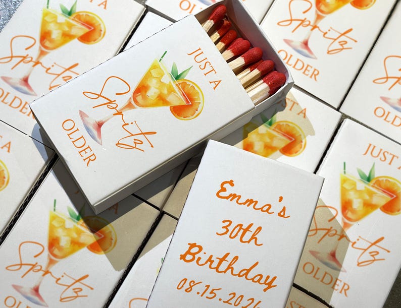 May include: White matchboxes with orange text and a watercolor illustration of a spritz cocktail. One box is open, revealing red-tipped matches. Text on the boxes reads "Just a Spritz Older" and "Emma's 30th Birthday 08.15.2026".
