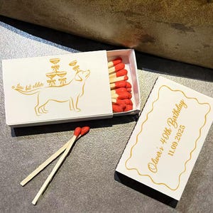 May include: White matchbox with a gold illustration of a dog and champagne glasses. The text "a fine bit older" is on the box. The matchbox is open, revealing red-tipped matches. A separate card reads "Oliver's 40th Birthday 11.09.2025".