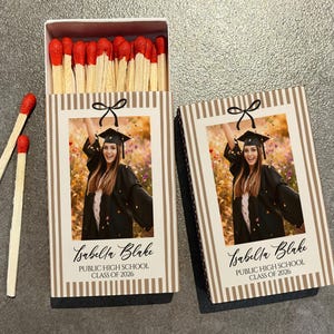 May include: Matchbox with a photo of a graduate in a cap and gown, with the text "Isabella Blake, Public High School, Class of 2026." The matchbox has a striped design with a black bow. Two matches are next to the box.