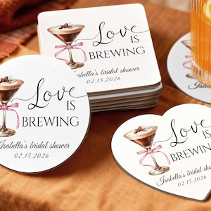 May include: White coasters with a watercolor illustration of a cocktail with a pink ribbon, coffee beans, and the text "Love is Brewing." The coasters are for Isabella's bridal shower on 02.15.2026. One coaster is heart-shaped.