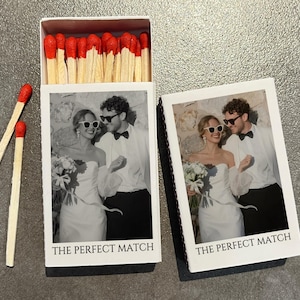 May include: A white matchbox with a black and white photo of a couple wearing sunglasses, with the text "THE PERFECT MATCH". The matchbox is open, revealing red-tipped matches. Two loose matches are next to the box.