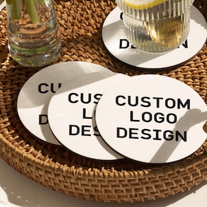 May include: A woven wicker tray holds several white circular coasters with the text "CUSTOM LOGO DESIGN" in black. A glass of water with a lemon slice and a clear glass vase are also on the tray.