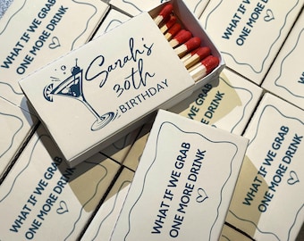 May include: A collection of matchboxes with a cream-colored background. The top matchbox is open, revealing red-tipped matches. The box is decorated with a blue cocktail graphic and the text "Sarah's 30th BIRTHDAY". Other boxes have the text "WHAT IF WE GRAB ONE MORE DRINK".