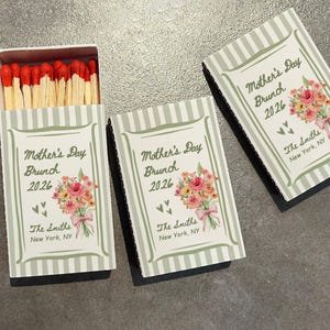 May include: Matchboxes with a Mother's Day Brunch 2026 theme. The boxes are white with green stripes and feature a floral bouquet and the text "The Smiths, New York, NY". The match heads are red.