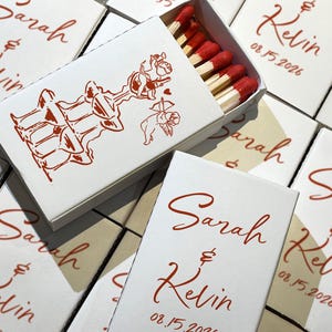 May include: White matchboxes with red text and illustrations. One box is open, revealing red-tipped matches. The boxes are personalized with the names "Sarah & Kelvin" and the date "08.15.2026". The boxes feature a decorative illustration.
