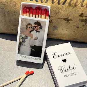 May include: A white matchbox with a photo of a couple and the text "THE PERFECT MATCH." The matchbox is open, revealing red-tipped matches. A second matchbox is open, with the names "Emma" and "Caleb," a heart, and the date "08.15.2026."