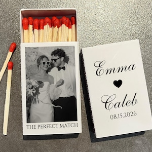 May include: A white matchbox with a black and white photo of a couple wearing sunglasses. The text "THE PERFECT MATCH" is below the photo. The matchbox is open, revealing red-tipped matches. The closed lid has the names "Emma" and "Caleb" with a heart symbol and the date "08.15.2026".