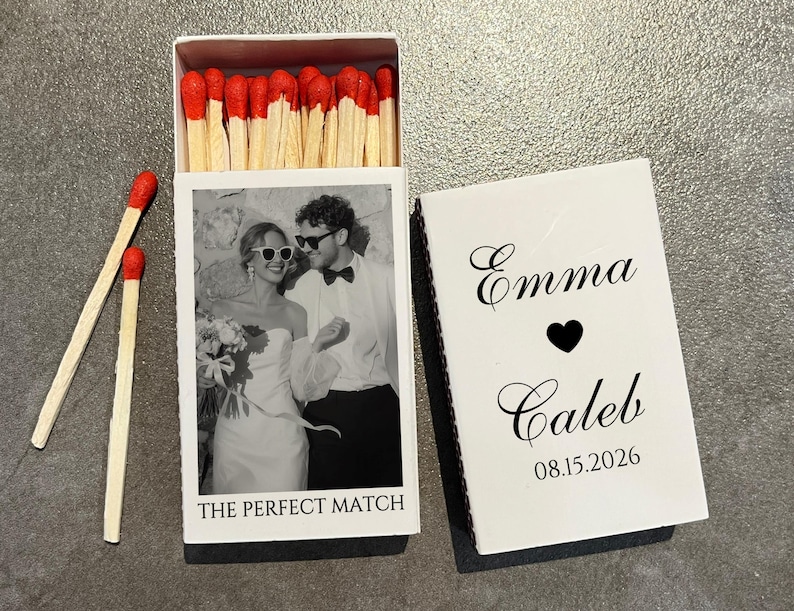 May include: A white matchbox with a black and white photo of a couple wearing sunglasses. The text "THE PERFECT MATCH" is below the photo. The matchbox is open, revealing red-tipped matches. The closed lid has the names "Emma" and "Caleb" with a heart symbol and the date "08.15.2026".