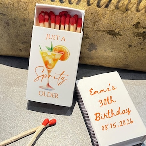 May include: White matchbox with "Just a Spritz OLDER" text and a cocktail illustration. The box is open, revealing red-tipped matches. A second box reads "Emma's 30th Birthday 08.15.2026".