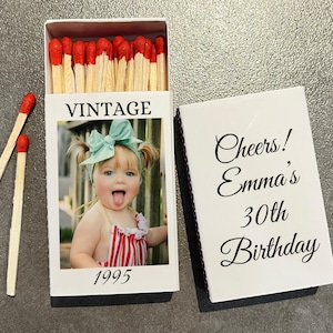 May include: A white matchbox with a photo of a baby girl with a mint green bow and red and white striped outfit. The box reads "VINTAGE" and "1995". The matchbox cover reads "Cheers! Emma's 30th Birthday". Two matches are next to the box.