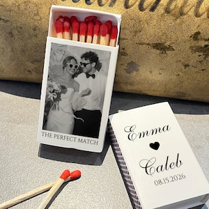 May include: A white matchbox with a black and white photo of a couple wearing sunglasses. The text "THE PERFECT MATCH" is below the photo. The matchbox is open, revealing red-tipped matches. A second matchbox is closed, with the names "Emma" and "Caleb", a heart, and the date "08.15.2026".