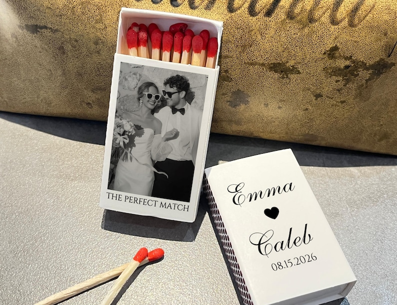 May include: A white matchbox with a black and white photo of a couple wearing sunglasses. The text "THE PERFECT MATCH" is below the photo. The matchbox is open, revealing red-tipped matches. A second matchbox is closed, with the names "Emma" and "Caleb", a heart, and the date "08.15.2026".