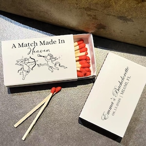 May include: White matchbox with the text "A Match Made in Heaven" and illustrations of cherubs. The box is open, revealing red-tipped matches. Two matches are placed in front of the box. A separate card reads "Emma's Bachelorette 08.15.2026 | MIAMI, FL."